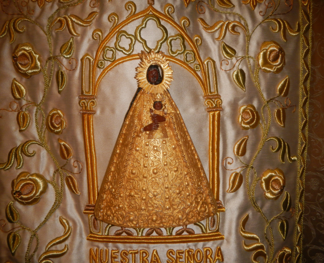 Goldbestickte Chasuble mit einer friedlichen Frau in einem hochgeschlossenen, langärmeligen Kleid mit einem Dutt, umgeben von detaillierten Designs und prächtigem Text in einem goldenen Rahmen.