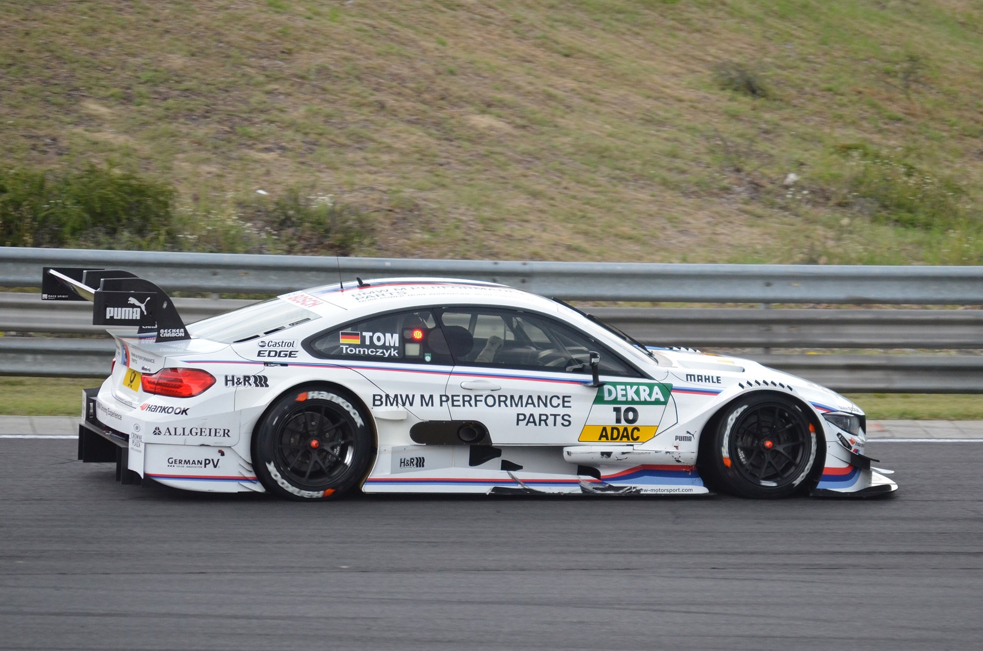 BMW M4 DTM Rennauto auf einer Rennstrecke mit Sponsor-Text, umgeben von grünem Gras und einer Metallschranke im Hintergrund.