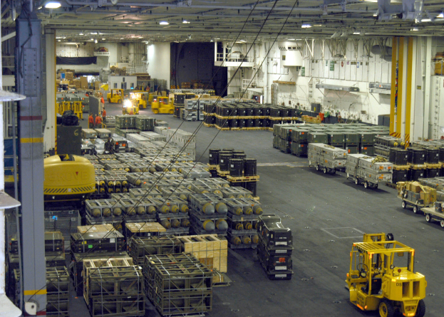 Ein großes Lagerinneres voller Lkw, Container, Menschen, Lampen, Stangen und ein deutliches Rad.