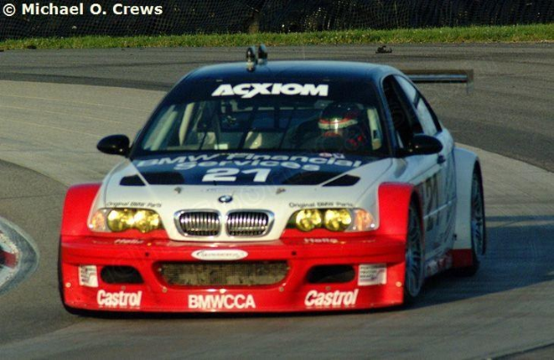 Eine Person in einem Helm fährt einen BMW M3 GT3 Rennwagen, der mit Text und Zahlen auf einer von Gras umgebenen Rennstrecke markiert ist, die von einem Maschendrahtzaun umgeben ist, mit einem Wasserzeichen in der oberen linken Ecke.