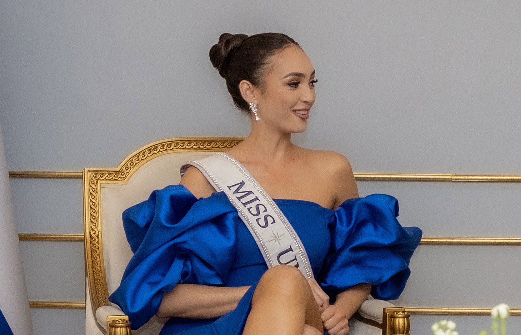 Eine Frau in einem blauen Kleid mit einer "Miss Universe 2019"-Schärpe sitzt lächelnd auf einem Stuhl umgeben von Blumen mit einer Wand im Hintergrund.