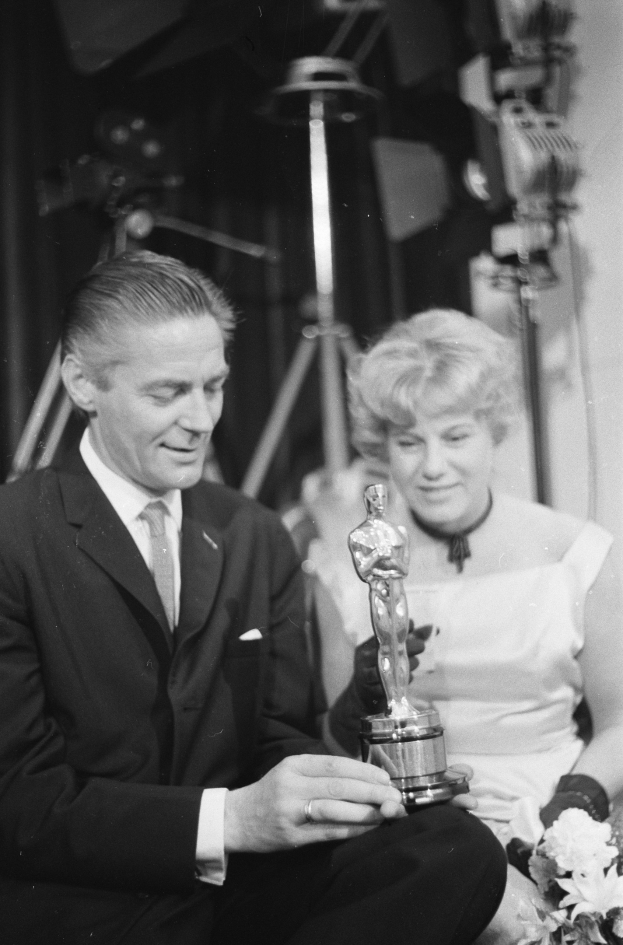 Ein Mann in einem schwarzen Blazer, weißem Hemd und Krawatte hält eine Oscar-Statue in der Hand und sitzt neben einer Frau, die einen Blumenstrauß hält, beide in eleganter Kleidung, vor einem schwach beleuchteten Hintergrund.