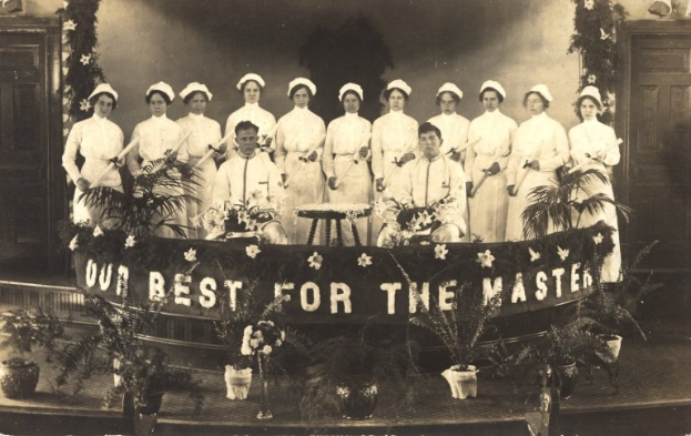 Eine Gruppe von Frauen in weißen Kleidern und Hauben steht auf einer Bühne mit einem Banner, auf dem 'Unser Bestes für den Meister' steht, umgeben von Blumentöpfen und mit Türen und einer Wand im Hintergrund.