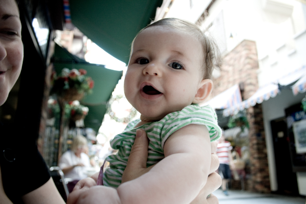 Eine Frau mit einem Kind auf dem Arm vor einem unscharfen Hintergrund mit einer sitzenden Frau, einem gehenden Mann, Hauspflanzen, Gebäuden und verschiedenen Gegenständen.