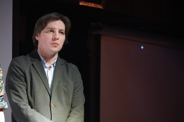 Zwei Personen stehen nebeneinander auf einer Bühne, eine trägt einen Anzug und die andere hat einen ernsten Gesichtsausdruck.