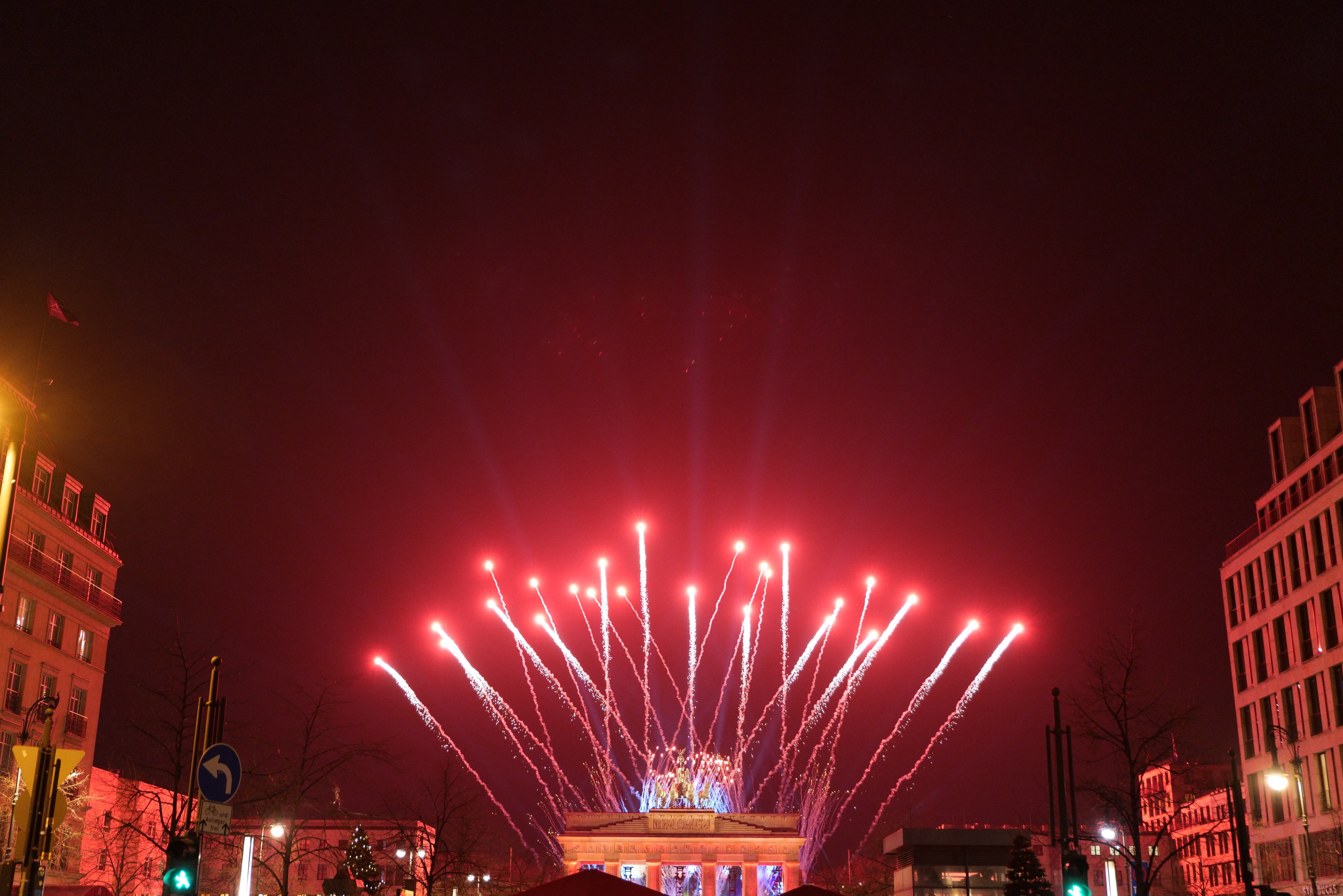 Eine nächtliche Stadtstraße in Berlin, Deutschland, mit dem Brandenburger Tor in der Mitte, umgeben von illuminierten Gebäuden, Bäumen, Ampeln, Schildern und Flaggen, und Feuerwerk, das den Himmel über dem Tor erhellt.