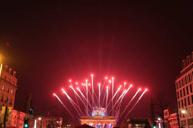 Eine nächtliche Stadtstraße in Berlin, Deutschland, mit dem Brandenburger Tor in der Mitte, umgeben von illuminierten Gebäuden, Bäumen, Ampeln, Schildern und Flaggen, und Feuerwerk, das den Himmel über dem Tor erhellt.