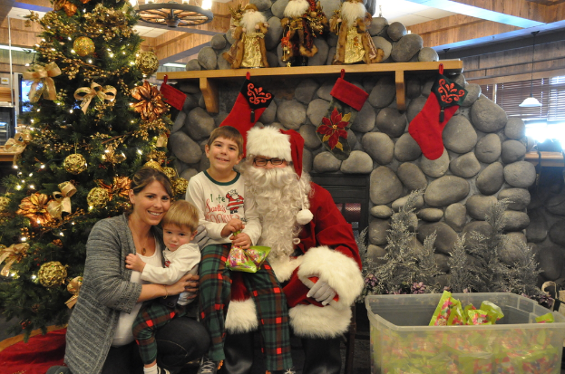 Eine Frau, Kinder und ein Mann als Santa Claus mit Brille und Handschuhen stehen neben einem geschmückten Weihnachtsbaum, einer Wand mit Felsen, Spielzeugen, Dekorationen und einem Korb mit Gegenständen, mit Lichtern im Hintergrund.