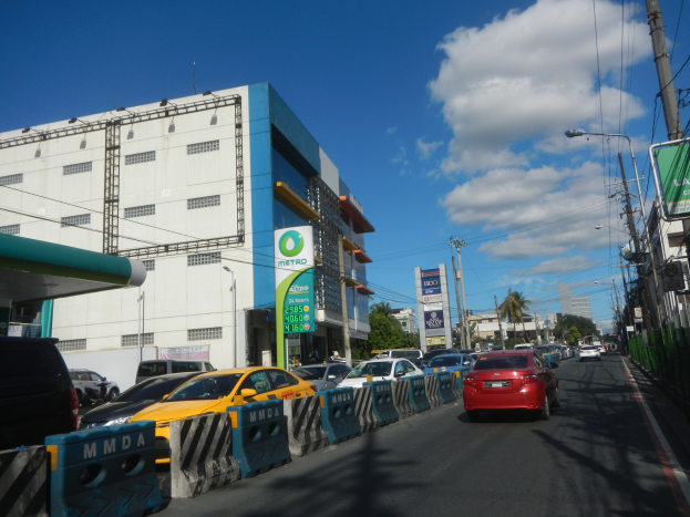 Eine belebte Stadtstraße mit starkem Verkehr, hohen Gebäuden, Absperrungen, Beschilderungen, Laternen, Strommasten, Bäumen und einem bewölkten Himmel.