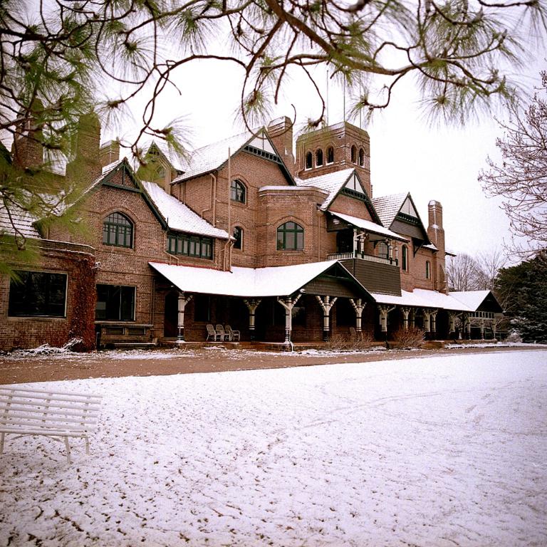 Ein Geb√§ude mit Fenstern, S√§ulen und St√ľhlen, mit einer Bank und einem Zweig auf der rechten Seite.