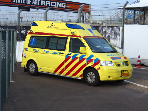 Ein Rettungswagen steht vor einem Zaun mit einem Verkehrskegel auf der rechten Seite der Straße, mit einem Bogen mit Text im Hintergrund und dem Himmel oben sichtbar.