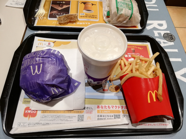 Ein Tisch mit einem Tablett, auf dem ein McDonald's-Essen mit Pommes, einem Kaffee, einem Blatt Papier, einer Gabel, einem Messer und anderen Gegenständen liegt.
