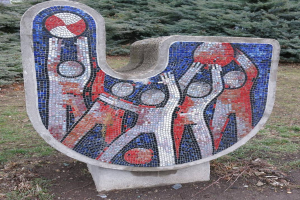 Eine Mosaikbank in einem Park, umgeben von Bäumen unter einem klaren blauen Himmel, mit farbigen Fliesen, die ein einzigartiges Design schaffen.