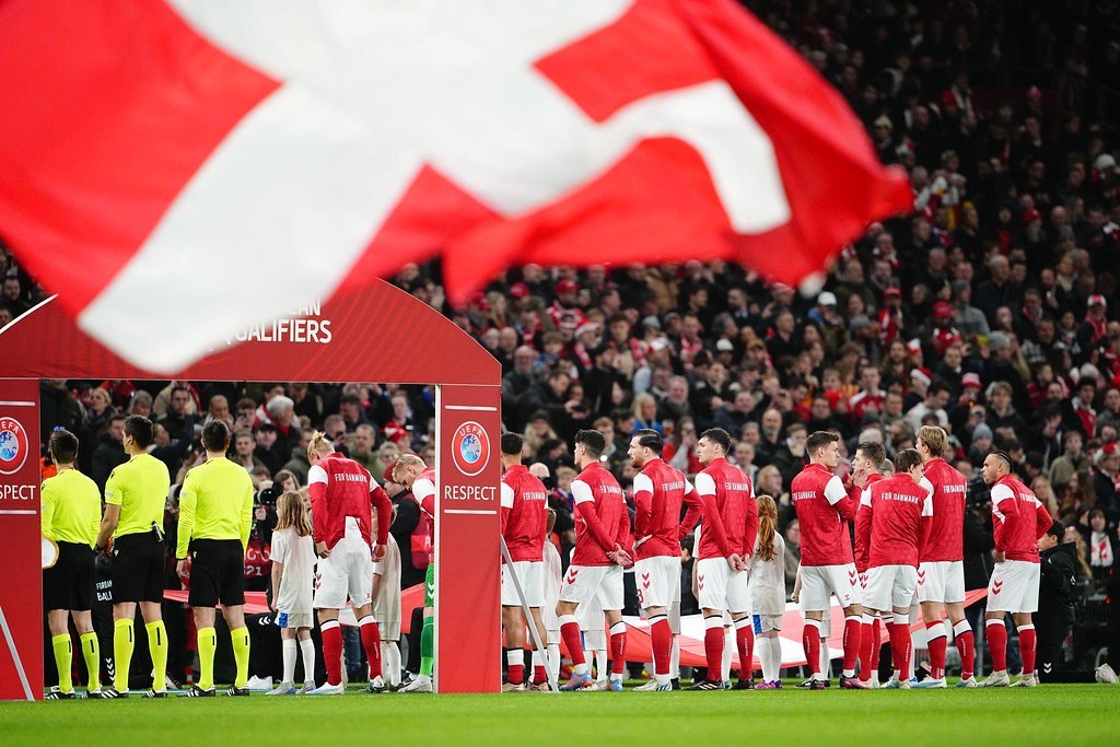 Gruppe von Menschen auf einem Fußballfeld mit einer roten und weißen Fahne im Vordergrund, einem Bogen mit der Aufschrift "Bayern München vs. Bayern München Wetten & Vorschau" im Hintergrund und einer großen Menge im Stadion.