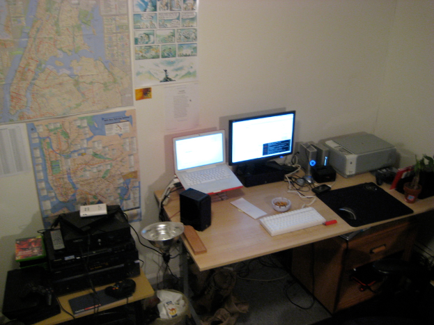 Ein Raum mit einem Tisch, auf dem ein Laptop, Lautsprecher, Monitor, Tastatur, Ball, Maus, Scanner und Papiere liegen, sowie ein weiterer Tisch mit Elektronikgeräten im Hintergrund, mit Charts und Postern an der Wand dahinter.