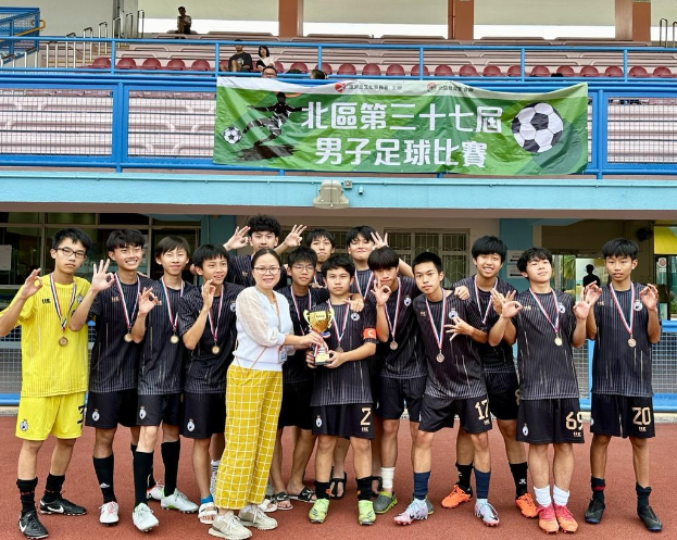 Gruppe junger Männer in Fußballtrikots auf einem Feld, die Medaillen tragen und einen Pokal halten, mit einem Banner im Hintergrund, das 'Yokohama U-16 Jungen Fußballmannschaft' zeigt, und Zuschauern in der Nähe sitzend.