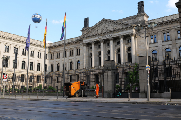 Großes modernes Gebäude mit zahlreichen Fenstern, als Bundestag beschriftet, umgeben von Straßeninfrastruktur, Fußgängern, Flaggen und Bäumen unter einem bewölkten Himmel.