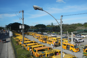Eine große Anzahl von gelben Schulbussen am Straßenrand geparkt, mit Menschen, die auf dem Gehweg gehen, Strommasten mit Drähten, Laternen, Bäumen, Gebäuden und einem bewölkten Himmel im Hintergrund.