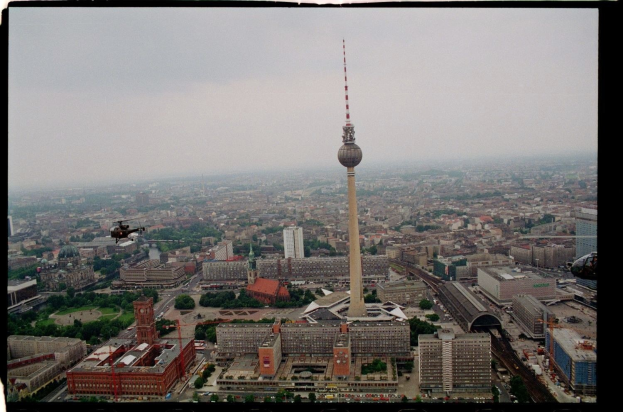 Luftaufnahme von Berlin, Deutschland, mit dem zentralen Fernsehturm, umgeben von Gebäuden, Bäumen, Straßen, Fahrzeugen und einem Hubschrauber unter einem klaren blauen Himmel.