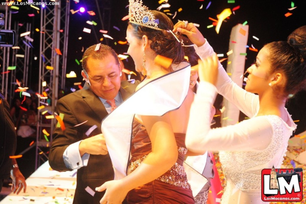 Ein Mädchen auf der Bühne bei einer Miss World Show mit einer Krone, dahinter ein Mädchen, das die Krone anpasst, und vorne ein Mann im schwarzen Anzug, der die Hand schüttelt, mit Metallrahmen und dunklem Hintergrund.