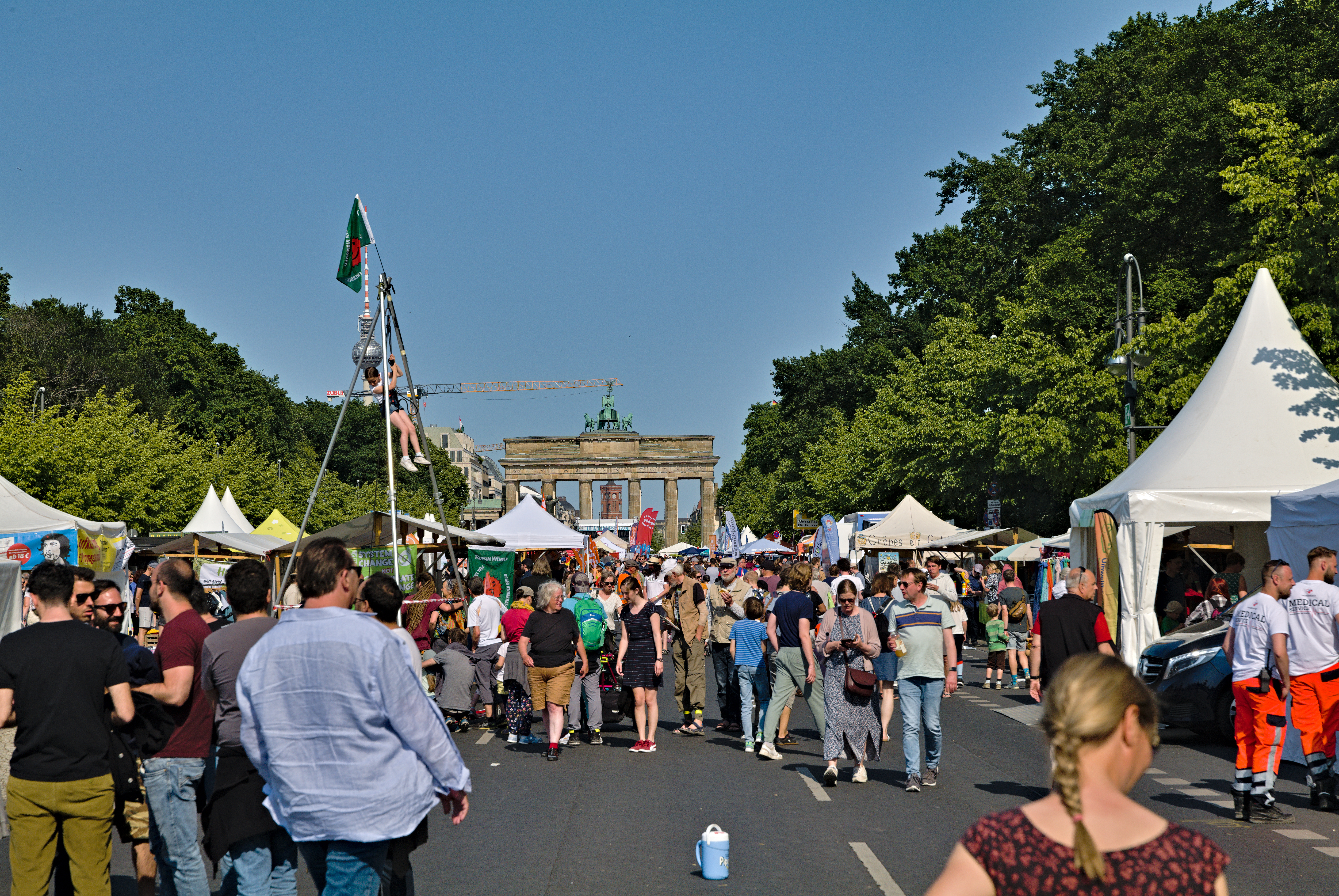 Eine Menschenmenge, die eine Straße mit Zelten, Fahrzeugen und Bäumen entlanggeht, mit einem Bogen und einem klaren blauen Himmel im Hintergrund und Masten mit Flaggen auf der linken Seite, wahrscheinlich das Oktoberfest in München, Deutschland darstellend.