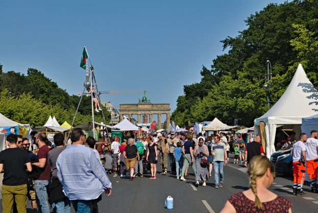 Eine Menschenmenge, die eine Straße mit Zelten, Fahrzeugen und Bäumen entlanggeht, mit einem Bogen und einem klaren blauen Himmel im Hintergrund und Masten mit Flaggen auf der linken Seite, wahrscheinlich das Oktoberfest in München, Deutschland darstellend.