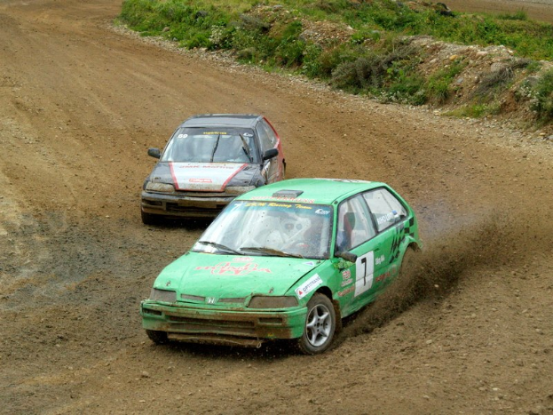 Zwei Autos mit Text und Zahlen fahren auf einer Schotterstraße, umgeben von Gras, Pflanzen und Steinen, was auf ein Rallye-Veranstaltung hinweist.