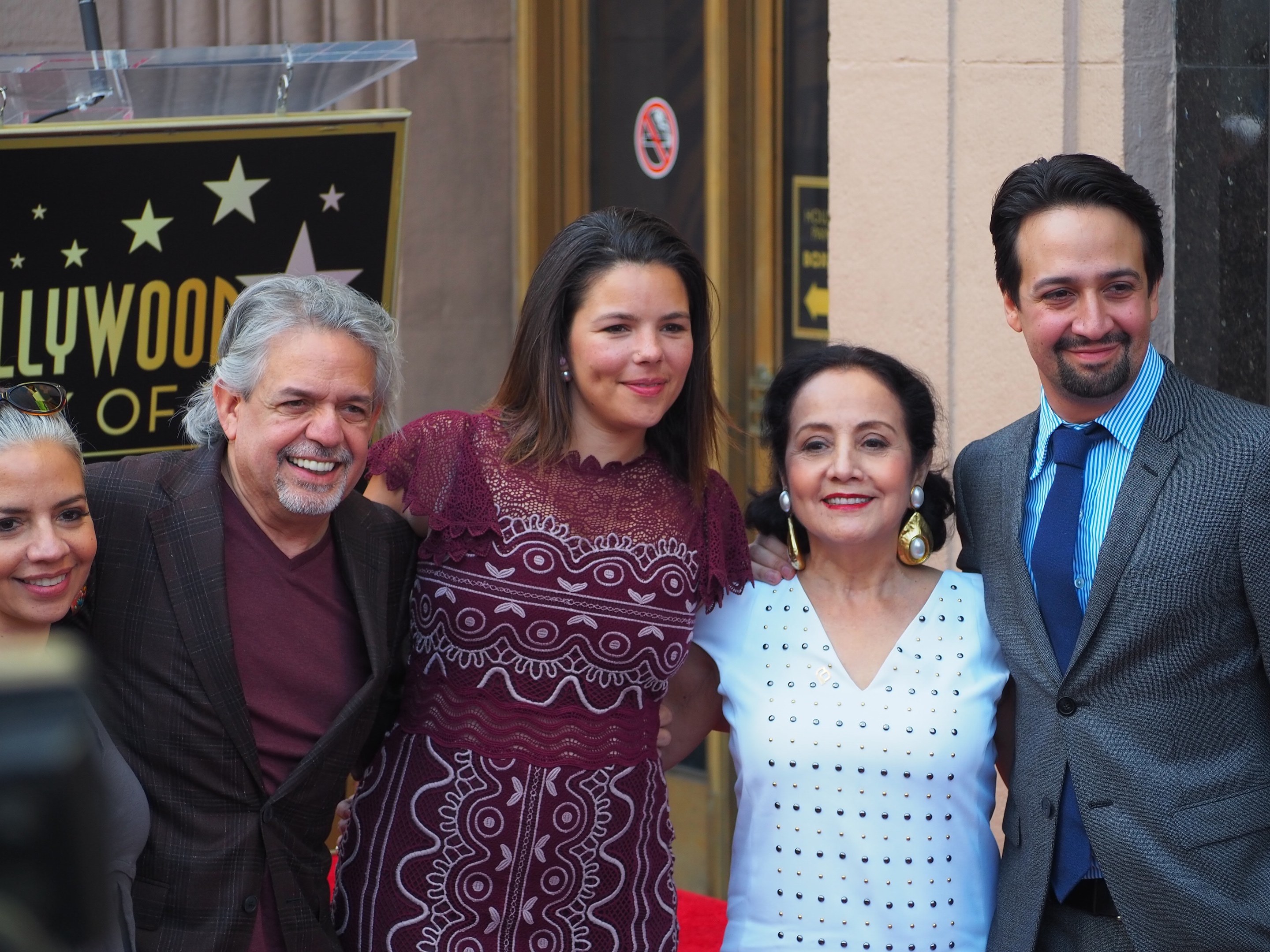 Gruppe von Menschen auf einem roten Teppich bei der Hollywood Walk of Fame Zeremonie mit einer Kamera auf der linken Seite, einer Tafel mit Text, einem Glaspodest und einer Wand im Hintergrund.