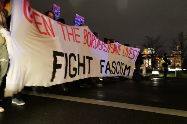 Eine Gruppe von Menschen marschiert mit einem Banner, auf dem "Kämpft gegen Faschismus" steht, durch die Straße, wobei einige Schilder halten und verschiedene Kleidung tragen, darunter Turnschuhe, unter einem klaren blauen Himmel mit Bäumen und Gebäuden im Hintergrund.