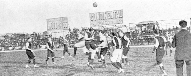 Ein Schwarz-Weiß-Bild einer Gruppe von Männern, die auf einem Feld Fußball spielen, mit einem Ball in der Luft, Zuschauern im Hintergrund und Tafeln mit Text, der wahrscheinlich Spielinformationen enthält.