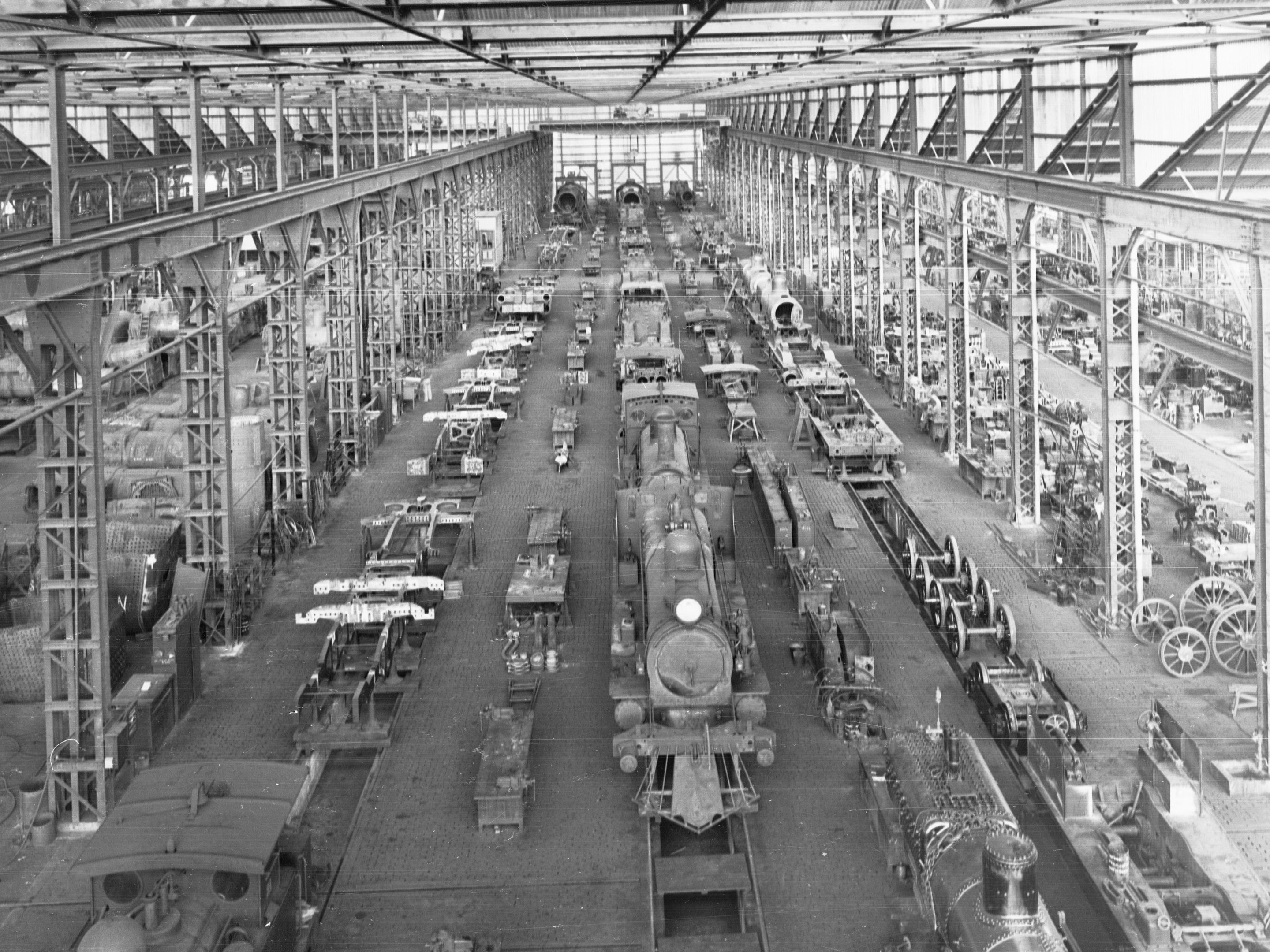 Schwarzes und weißes Foto einer großen Fabrikhalle mit zahlreichen Zügen, Fahrzeugen und Gegenständen auf dem Boden, Metallstangen und einer Decke.