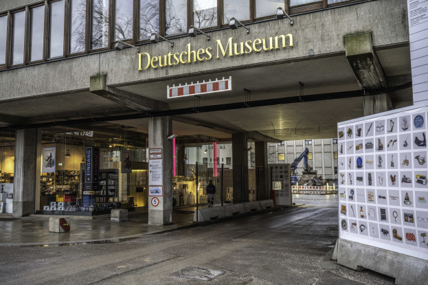Außensicht des Deutschen Museums in Berlin: Ein großer Bau mit Glasfenstern, Säulen und einem Namensschild, neben einer Informationsanzeigetafel, mit anderen Gebäuden, Bäumen und Himmel im Hintergrund.