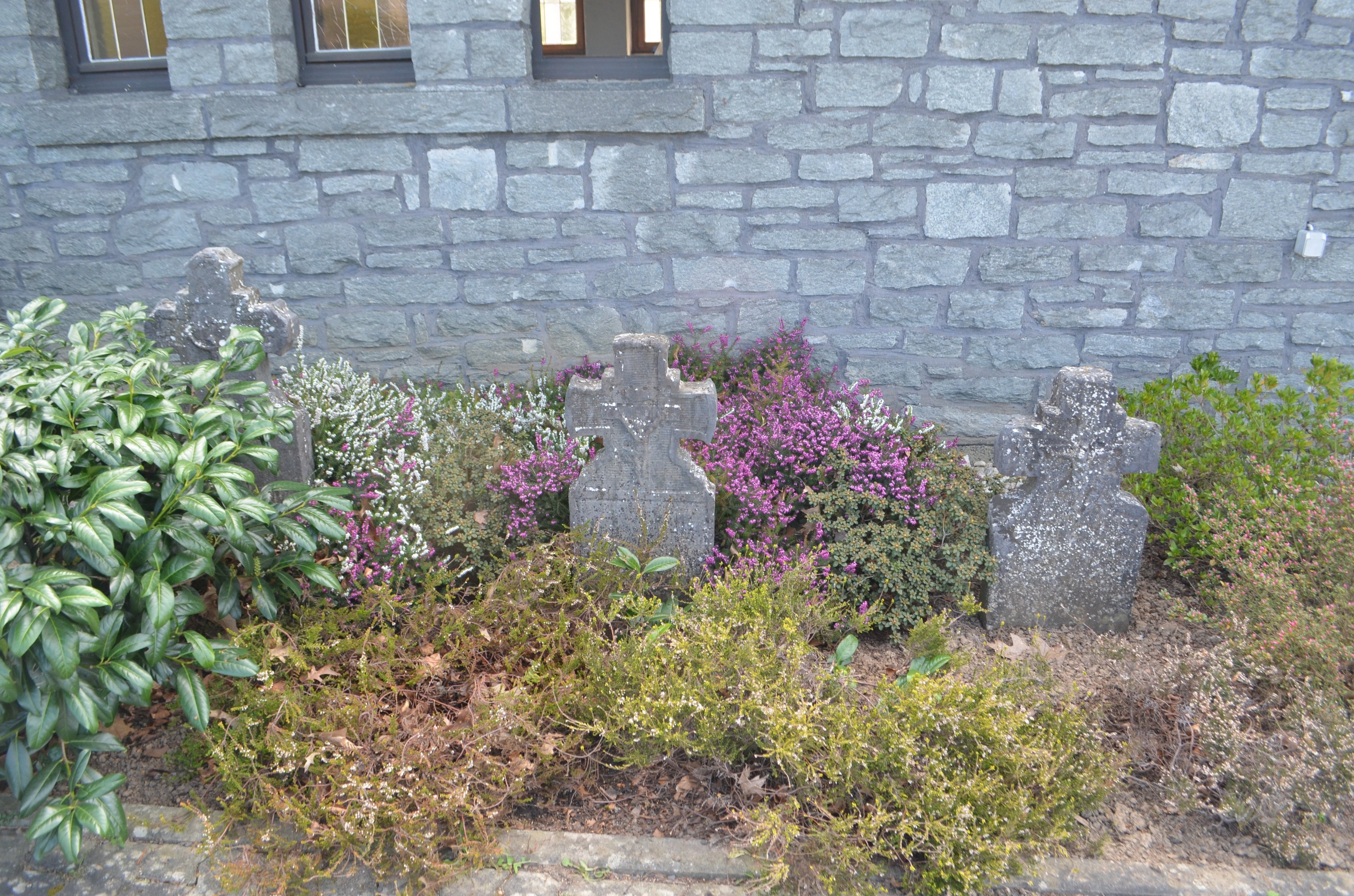 Steinbau mit Garten, der Pflanzen, Blumen und Grabsteine vor dem Gebäude zeigt, Fenster im Gebäude sichtbar