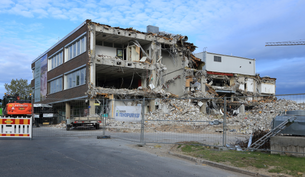 Ein abgerissenes Gebäude, umgeben von einem Metallzaun, mit Fahrzeugen, Gras, einem Schild, einer Fahne, Pfählen, einem Kran, Bäumen und einem bewölkten Himmel.