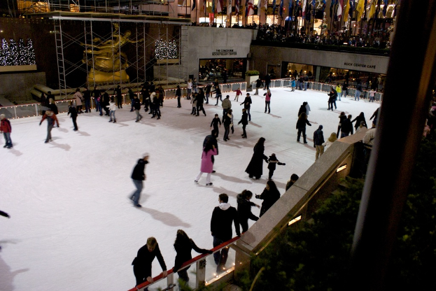 Eine Gruppe von Menschen, die auf einer zugefrorenen Oberfläche Schlittschuh laufen, umgeben von Gebäuden.