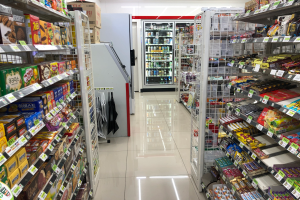 Ein Supermarktgang voller verschiedener Lebensmittel auf Regalen mit Preisschildern, ein Kühlschrank im Hintergrund und Deckenleuchten.