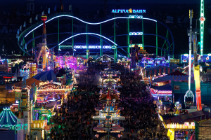 Eine große Menschenmenge versammelt sich um einen nächtlichen Karneval, der von bunten Lichtern beleuchtet wird, mit Vergnügungsgeräten, Pfählen, Fahnen und Namensschildern im Hintergrund.