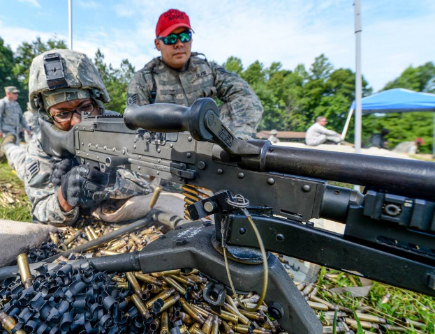 Eine Person auf einem Armee-Trainingsgelände feuert eine Waffe ab, während eine andere Person mit einer roten Mütze daneben steht, während im Hintergrund Bäume, Zelte und Menschen zu sehen sind und Patronenhülsen und Gras im Vordergrund liegen.