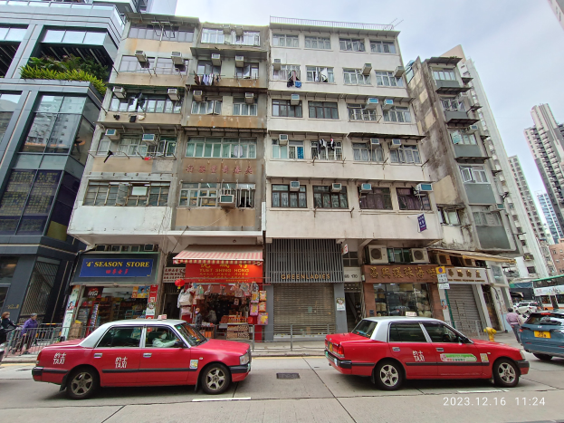 Rotes Taxi fährt eine Straße mit hohen Gebäuden, Fußgängern und verschiedenen Schaufenstern im Hintergrund.