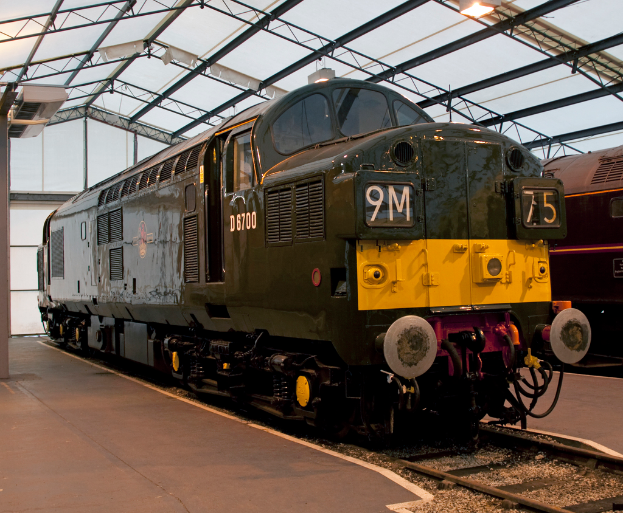 Ein Zug steht auf den Schienen neben einer Plattform mit einem an der Decke befestigten Mast, der Metallstangen mit Lampen trägt.