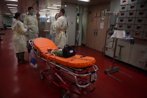 Eine Gruppe von Menschen in Schutzausrüstung steht um eine Trage in einem Krankenhauszimmer, das mit medizinischer Ausrüstung gefüllt ist, darunter eine Trage mit einer Tasche und anderen Gegenständen, ein Ständer mit verschiedenen Gegenständen, eine Tür, eine Säule, ein Schild und eine Decke mit Lampen.