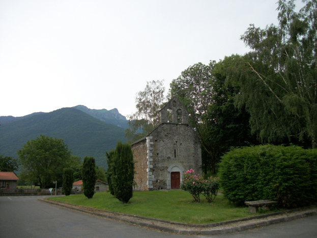Alte Kirche in der Mitte einer Straße, umgeben von Häusern, Grün, einer Bank und einer Straße mit Hügeln und einem blauen Himmel im Hintergrund.