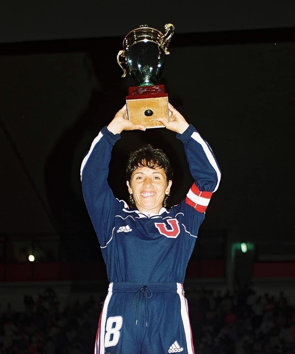 Eine Frau in einem blauen Kleid hält einen Pokal hoch und lächelt, umgeben von einer jubelnden Menge in einem hell erleuchteten Stadion.