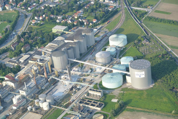 Luftaufnahme mit mehreren Geb√§uden, H√§usern, B√§umen und Fahrzeugen auf einer von Gras umgebenen Stra√üe.