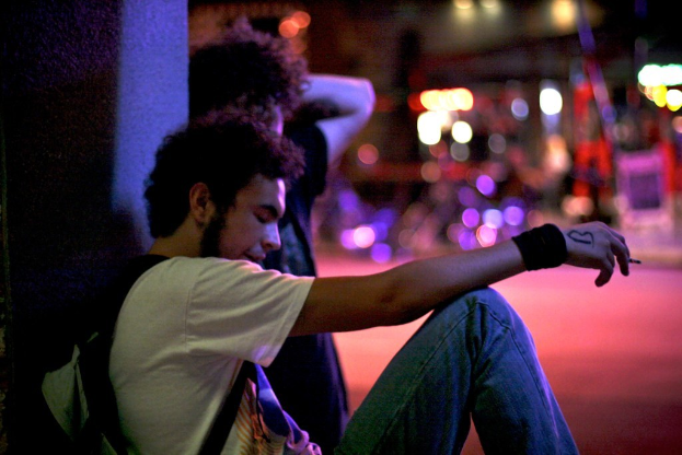 Mehrere Personen sitzen auf dem Boden, wobei eine Person eine Zigarette und eine Tasche in den Händen hält.