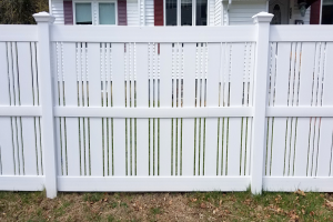 Ein weißer Vinyl-Privatszaun steht vor einem Haus mit Fenstern, umgeben von Gras und trockenen Blättern, mit Bäumen im Hintergrund.