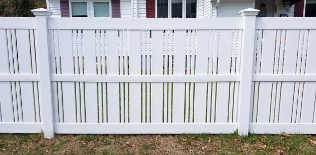 Ein weißer Vinyl-Privatszaun steht vor einem Haus mit Fenstern, umgeben von Gras und trockenen Blättern, mit Bäumen im Hintergrund.