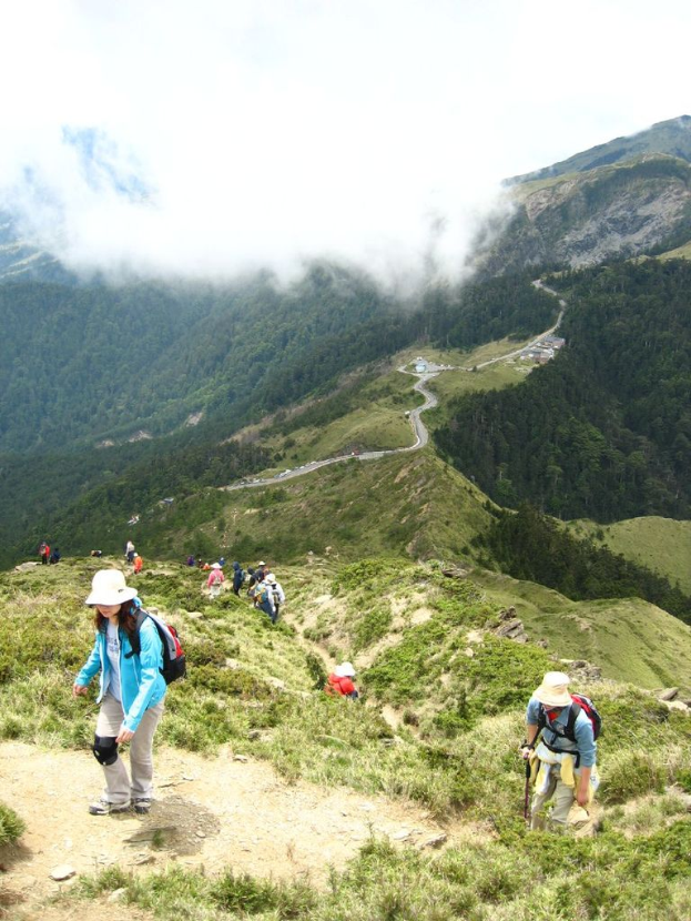 Eine Gruppe von Menschen, die einen Berg mit Bäumen und einer Straße hinaufsteigen, unter einem Himmel mit Wolken.