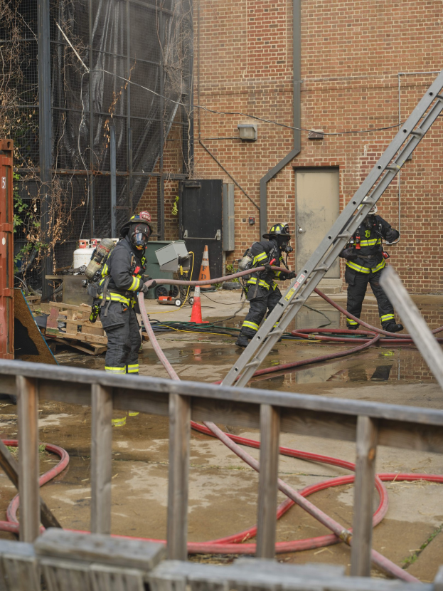 Feuerwehrleute mit Helmen arbeiten daran, ein Gebäude Feuer zu löschen, während sie in der Nähe eines Metallzauns, Rohren, eines Containers und eines Verkehrskegels stehen, mit einem Baum und Himmel im Hintergrund.