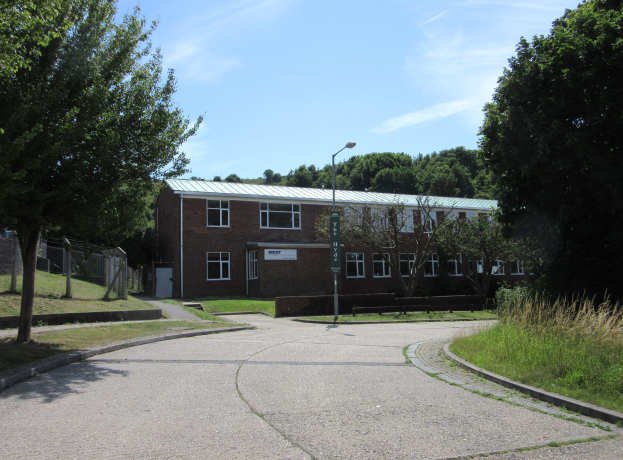 Ein Schulgebäude mit Fenstern, einem Straßenschild, einem Schild, einer Baumgruppe, Gras, einem Zaun und einem bewölkten Himmel, das in der Mitte eines Parks umgeben von saftigem Grün steht.